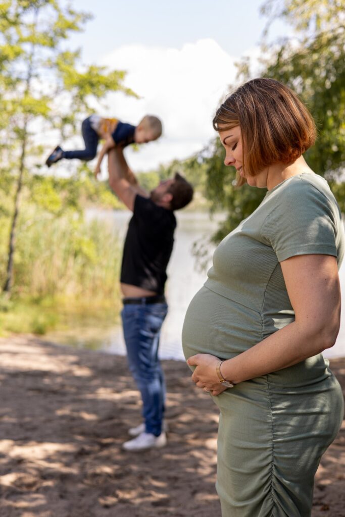 Papa hält Sohn in die Luft, Mama steht weiter vorne und schaut auf ihren Babybauch