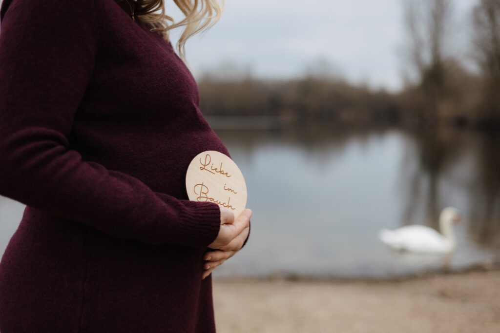 Schwangere Frau steht am See, hält ihren Bauch fest. Im Hintergrund schwimmt ein Schwan. - Detailfoto