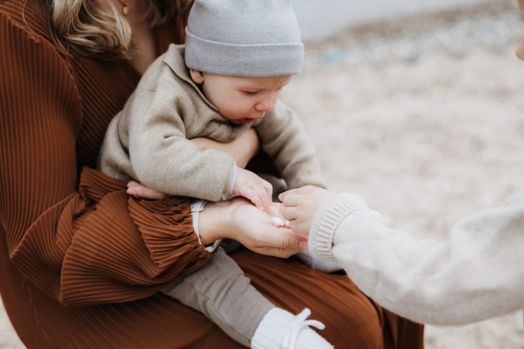 Mama hält ihr Baby auf dem Schoß und der große Bruder bringt Muscheln