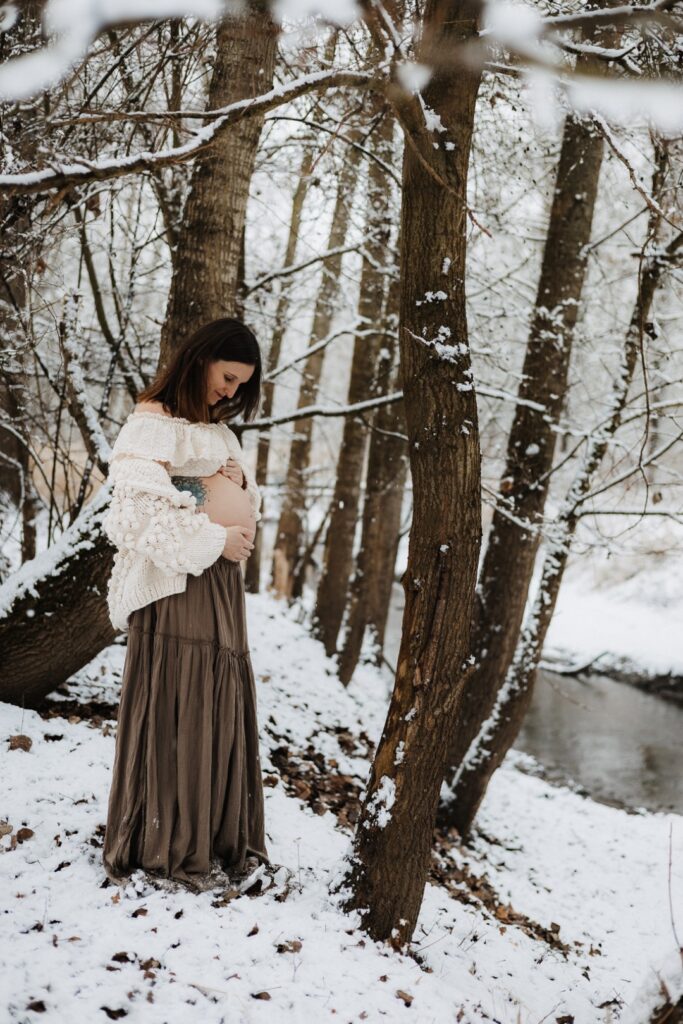 Schwangere Frau steht im Wald mit Schnee und schaut ihren Babybauch an
