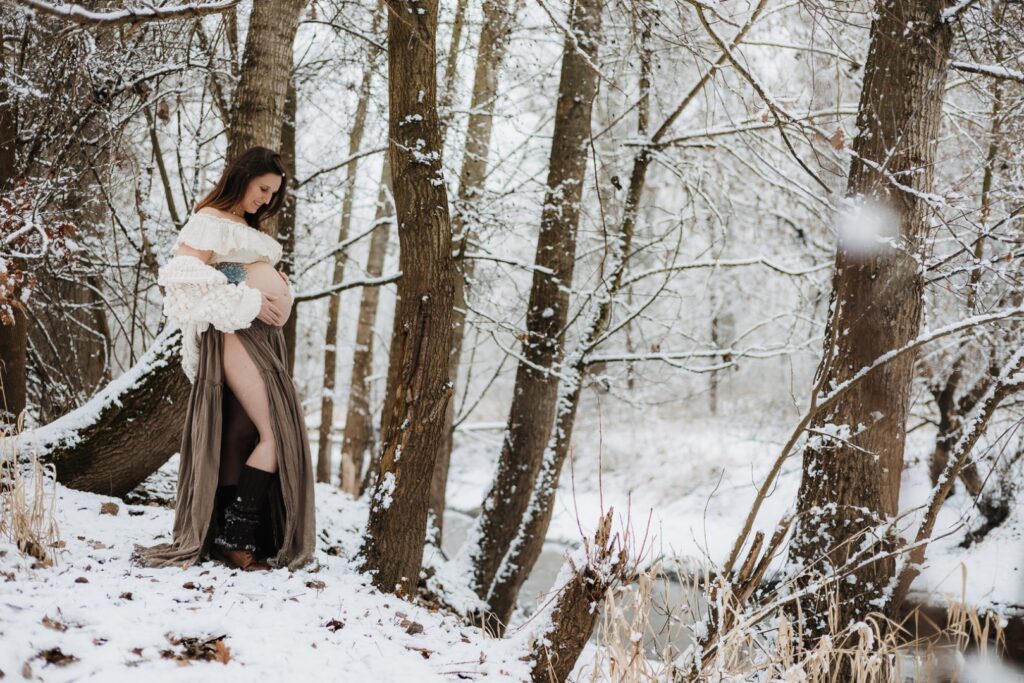 Schwangere Frau steht im Wald mit Schnee und schaut ihren Babybauch an