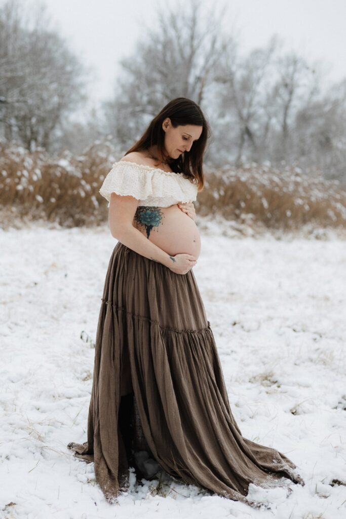 Schwangere Frau steht im Schnee mit langem braunen Rock und kurzer Bluse und schaut ihren Babybauch an