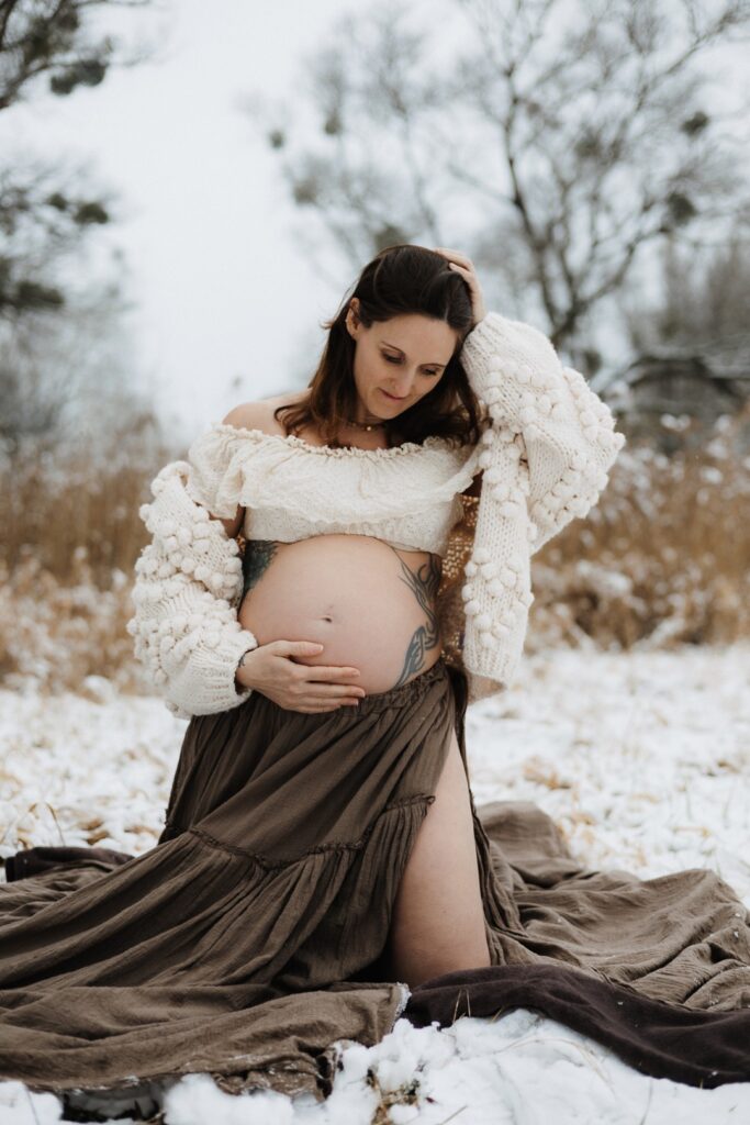 Schwangere Frau kniet auf einer Decke im Schnee und schaut ihren Babybauch an