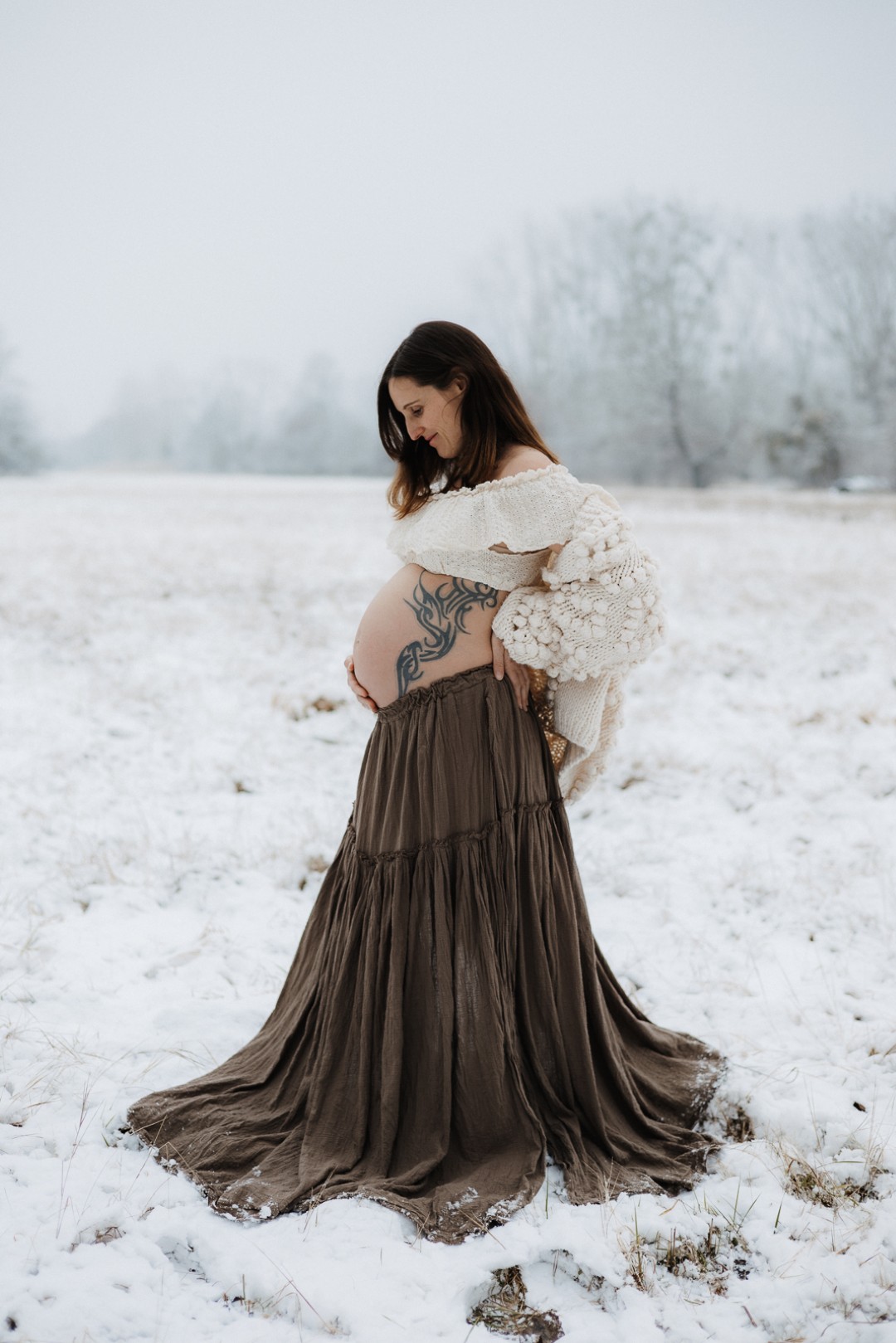 Schwangere Frau steht im Schnee mit langem braunen Rock und kurzer Bluse und schaut ihren Babybauch an