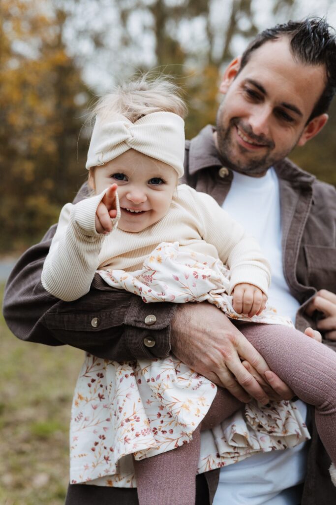Papa hat seine kleine Tochter auf dem Arm, sie lacht und zeigt mit dem Finger in die Kamera