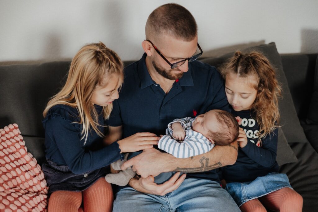 Papa hält seinen Sohn im Arm, zwei große Schwestern sitzen direkt danaben