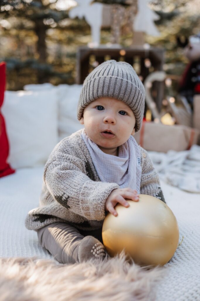 Kleinkind sitzt auf einer Decke im Tannenwald und spielt mit einer Christbaumkugel