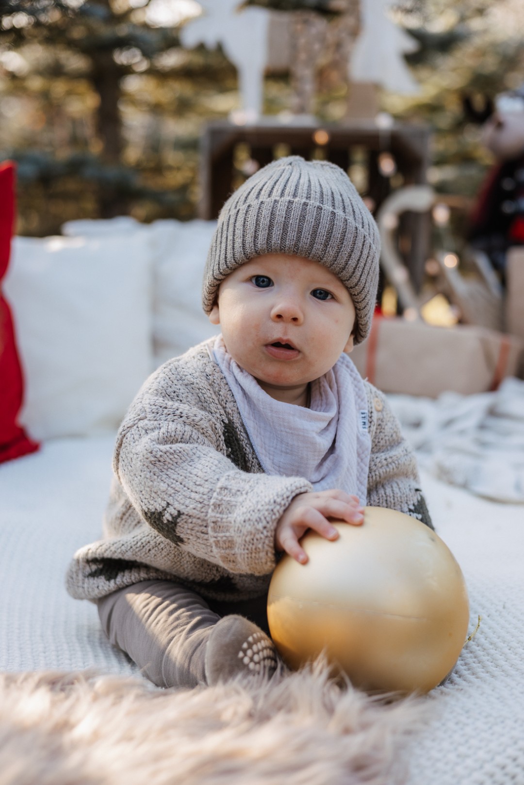 Kleinkind sitzt auf einer Decke im Tannenwald und spielt mit einer Christbaumkugel